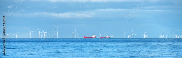 Fototapeta A vast expanse of ocean stretches under a bright blue sky, dotted with white wind turbines gently rotating in the distance. Two ships travel across the calm sea, adding a touch of vibrancy.