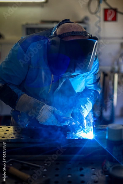 Fototapeta Man with welding mask in safety suit with gloves welds steel in home workshop. Welding flame with sparks. Dangerous work