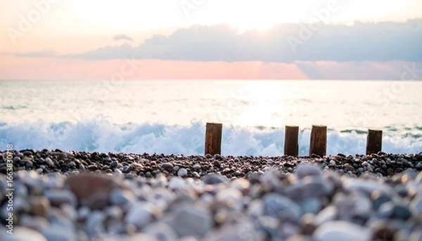 Fototapeta Wooden posts stand sentinel on a pebbled beach,  facing a serene ocean at sunset.