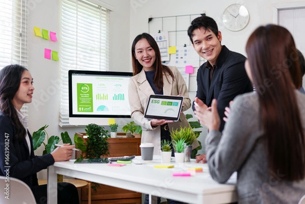 Fototapeta Young professionals in modern office discuss ESG data, smiling and collaborating around table with digital tablet and charts, surrounded by plants and sticky notes, teamwork atmosphere