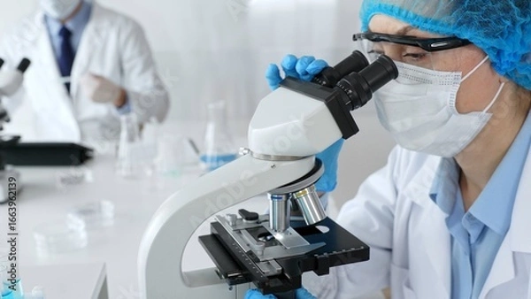 Fototapeta Female scientist wearing a mask and blue safety gear, using a microscope for research while a male colleague works in the background. Medicine, healthcare and science concept
