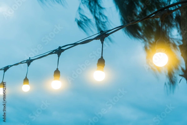 Obraz A lone bright light bulb hangs from a tree, illuminating a blue sky with soft, sunny clouds on a bright, beautiful summer day