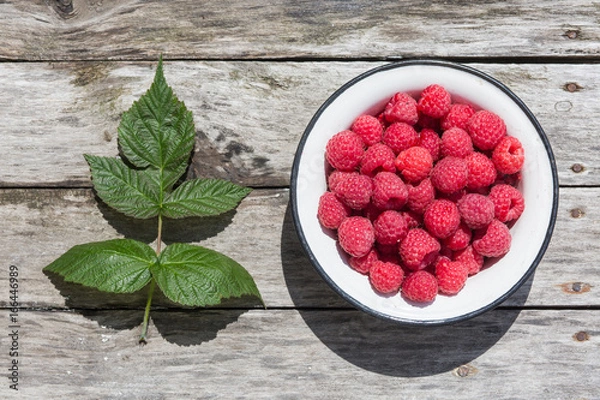 Fototapeta Raspberry in a white plate on an aged wood background. Green raspberry leaves.