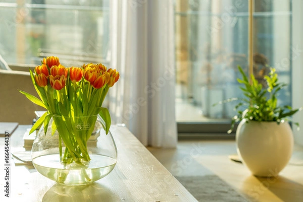 Fototapeta Tulips on a table in Sunlit Room: A bouquet of vibrant orange tulips is elegantly arranged in a glass vase on a wooden table. 