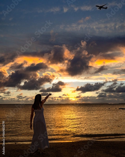 Obraz 南国のビーチで夕暮れ時に飛行機を指さすワンピース姿の女性のイメージ写真