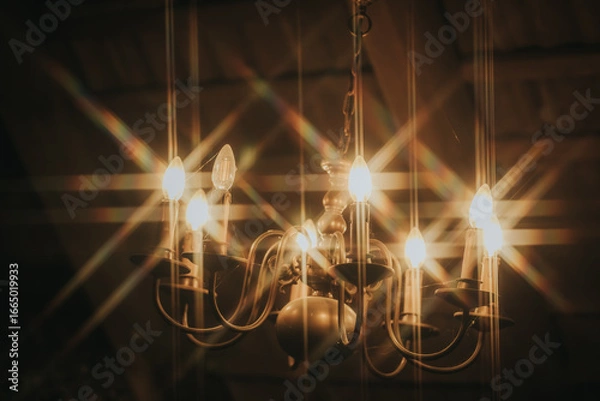 Fototapeta A vintage chandelier glows warmly, casting star-shaped light flares and adding charm to a cozy, rustic setting.
