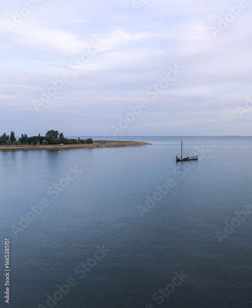Fototapeta A Tjalk a historic small sailboat floats gracefully in calm waters, close to the shore. The landscape features a serene coastline dotted with trees, against a softly clouded evening sky.

