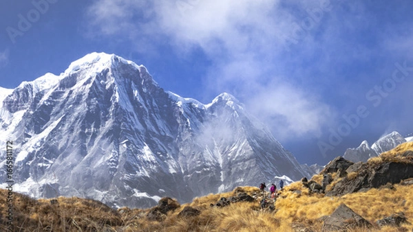 Fototapeta Trekkers trekking on rugged path towards Mt. Annapurna south mountain peak of nepal Himalaya under partial cloud cover. Grassland meadow covered in dry grass in foreground.