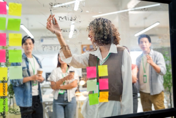 Fototapeta Asian business creative marketing team brainstorming startup strategy using glassboard and sticky notes. Collaborative ideation session demonstrates design thinking methodology disruptive innovation