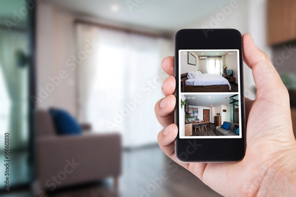 Fototapeta Hand holding smartphone view looking in house on blurred house background with Home security CCTV system concept.