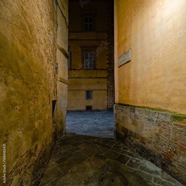 Fototapeta Narrow via del Poggio in Siena, illuminated by streetlights, leading to via San Pietro. historic architecture, stone-paved path, and ambiance create a  nighttime scene. Travel to Tuscany, Italy.