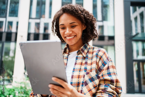 Fototapeta A joyful woman smiles brightly while using a tablet outdoors in a vibrant urban setting