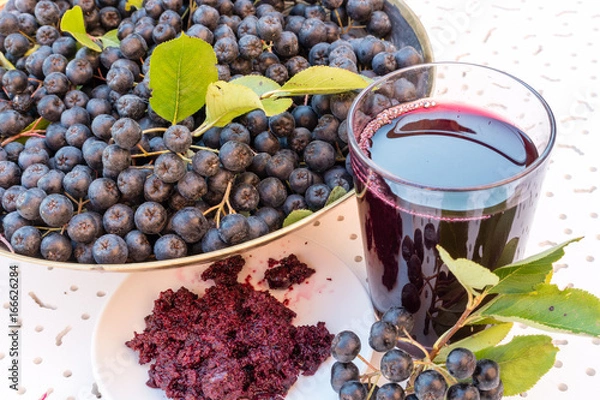 Fototapeta Closeup of fresh juice and jam of ripe black chokeberry (Aronia melanocarpa) in glass and berry in pot on white textured background