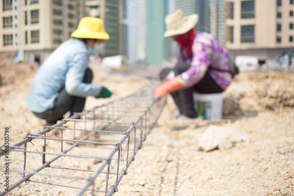 Fototapeta Asian migrant construction worker tying rebar framework amidst a cityscape of skyscrapers.