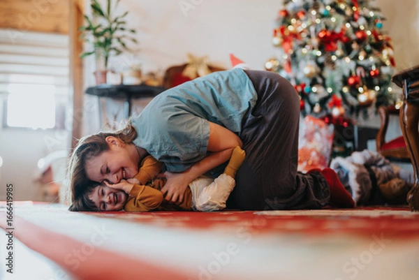 Fototapeta A mother and child embrace and laugh together in front of a beautifully decorated Christmas tree, capturing the festive spirit and joy of the holiday season.