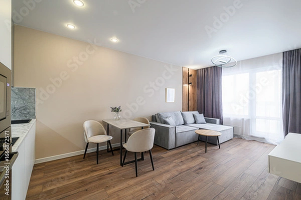 Fototapeta cozy apartment interior with a gray sofa, dining table, and warm wood flooring. Natural light fills the space