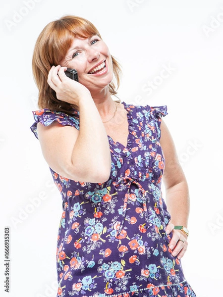 Fototapeta Phone Conversation. A mature woman talking on her mobile phone with a wide smile on her face. She is looking to the camera with a happy expression and wearing a purple floral dress.