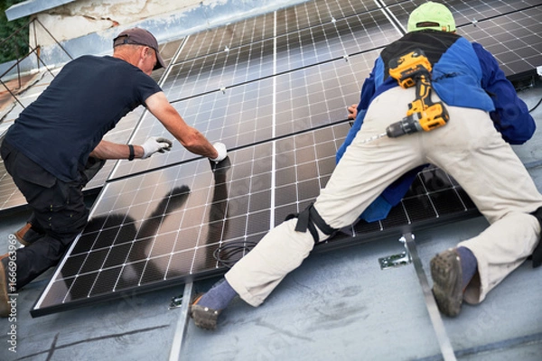 Fototapeta Workers building solar panel system on metal rooftop of house. Two men installers installing photovoltaic solar module outdoors. Alternative, green and renewable energy generation concept.