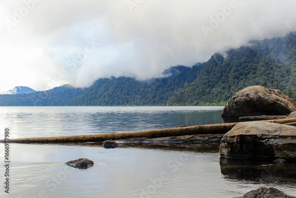Fototapeta Tranquil Lake Landscape with Misty Forest Mountains and Rocks in the Foreground