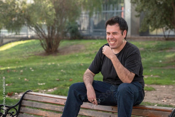 Fototapeta young man smiling and sitting on bench in park