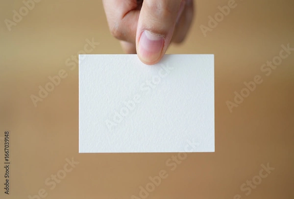 Fototapeta Hand holding a blank white square card against a textured brown background