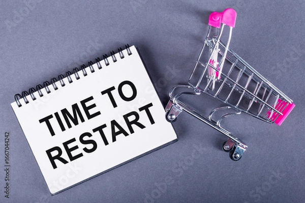 Fototapeta A notepad with the words TIME TO RESTART beside a miniature shopping cart on a gray background.