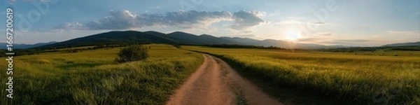 Fototapeta Serene pathway through lush green field at sunset with majestic mountain views