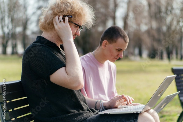 Fototapeta Three cheerful students study and work together outdoors on a sunny day, using laptops and notebooks while sitting on a park bench