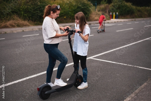 Fototapeta Two individuals engage with an electric scooter on outdoor pavement while one tries it out. Both seem actively immersed in learning and experimenting with the scooter