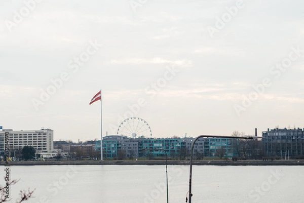 Fototapeta A calm riverside cityscape featuring a Latvian flag, a Ferris wheel, and modern buildings under a soft, overcast sky