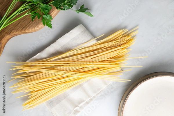 Fototapeta Uncooked spaghetti pasta on a linen cloth with fresh parsley on a wooden cutting board and an empty ceramic plate