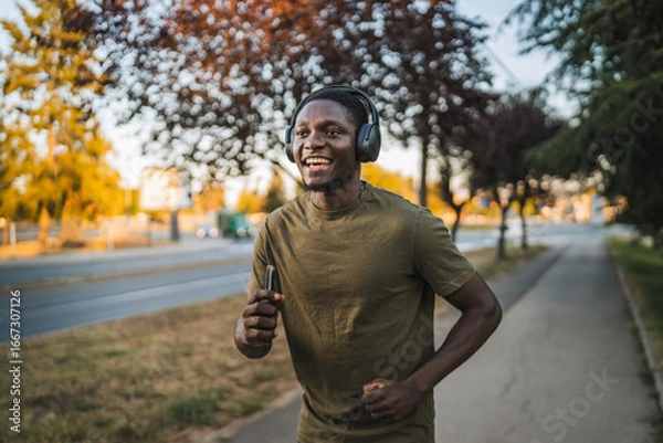 Obraz Young man running outdoors while listening to music with headphones and holding a smart phone