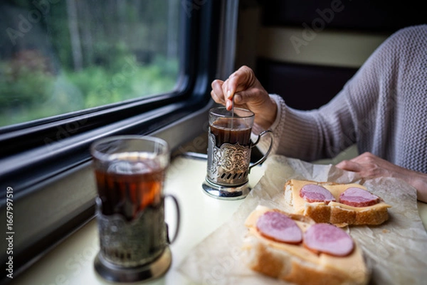 Fototapeta Passenger on train eating sandwich, drinking tea while look out window. Solo travel, railway commute, female passenger rest in train car on lunch break, slow living, journey transit, onboard dining