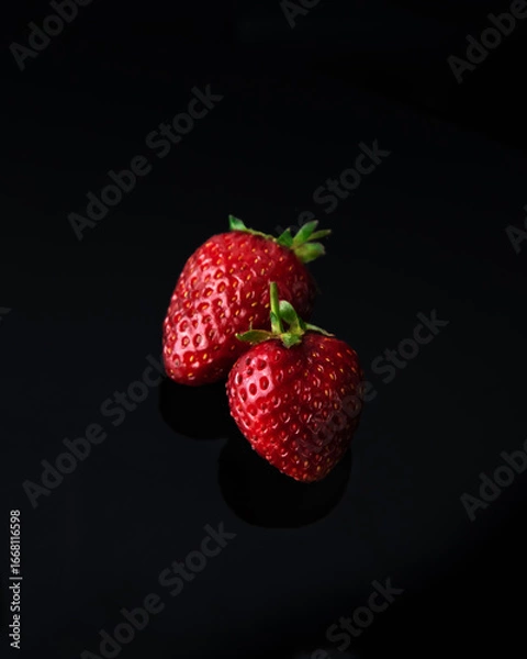 Fototapeta Close-up of two fresh strawberries with green stems on a reflective black surface. The red strawberries stand out against the dark background, highlighting their natural texture and vibrant color.