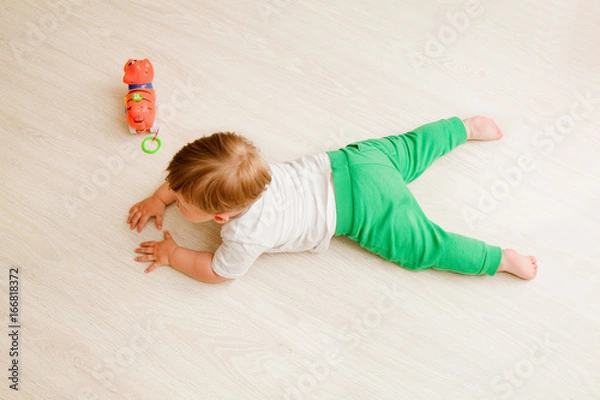 Fototapeta toddler boy white t-shirt and green pants lies on his stomach on the wooden floor. the view from the top
