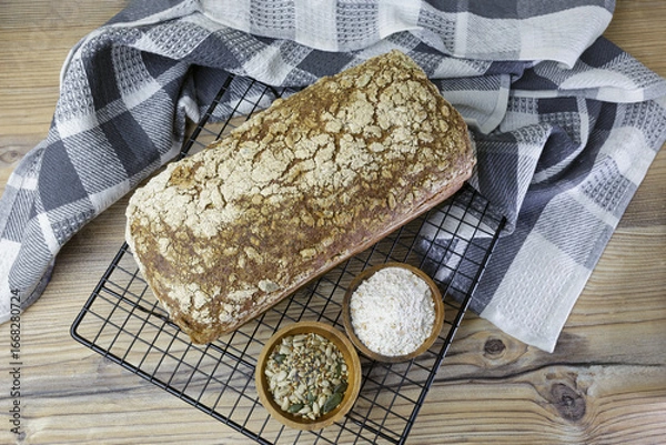 Fototapeta Flat lay of a loaf of bread resting on a cooling rack next to two bowls, one with flour and the other with sunflower seeds. The bread, made with whole-grain flour and healthy, is on a wooden table.