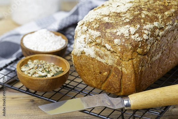 Fototapeta A loaf of bread rests on a wire rack next to a bowl of flour and another of seeds and a knife. The bread is whole wheat and has a crunchy texture.