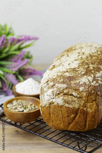 Fototapeta A loaf of bread rests on a wire rack next to a bowl of flour and another of seeds. The bread is whole wheat and has a crunchy texture. The simple scene has a sprig of lavender in the background.