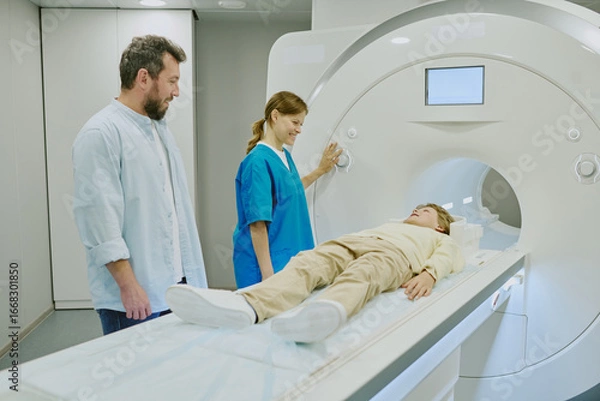 Fototapeta Caucasian boy lying on MRI scanner bed preparing for medical imaging procedure, young Caucasian woman technician operating machine, middle aged Caucasian man standing nearby observing