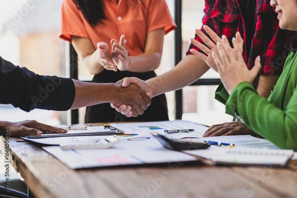 Fototapeta Business partners shaking hands at a meeting while their coworkers are clapping, celebrating the closing of a deal or a successful agreement.