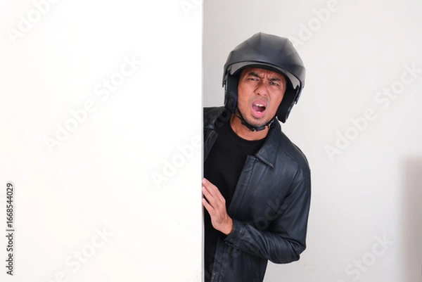 Fototapeta Motorcycle rider wearing helmet peeking from behind wall showing disappointed expression