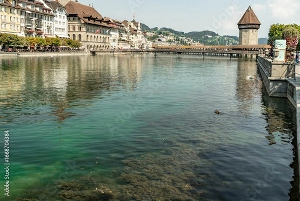 Fototapeta Chapel Bridge - a wooden footbridge in the city of Lucerne in Switzerland with a water tower in a sunny day, from the perspective of the Reuss river.