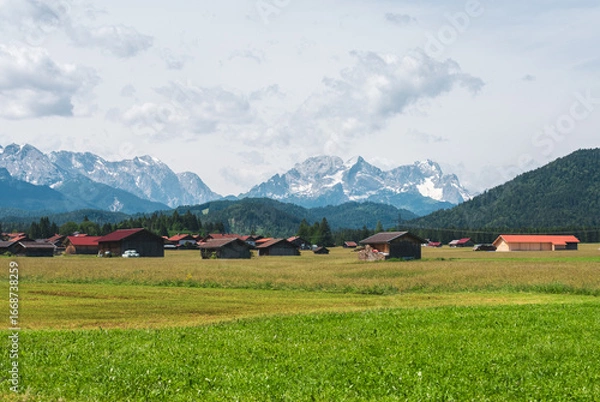 Obraz Traditional Bavarian barns and red-roofed farm buildings in green fields, with the snowy Alps rising in the background. A peaceful rural landscape in southern Germany.