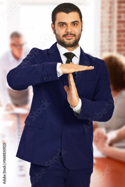 Obraz Confident businessman in a suit making a timeout gesture during a group office meeting, symbolizing the need for a break or pause. In the background, colleagues are engaged in discussion at the table.
