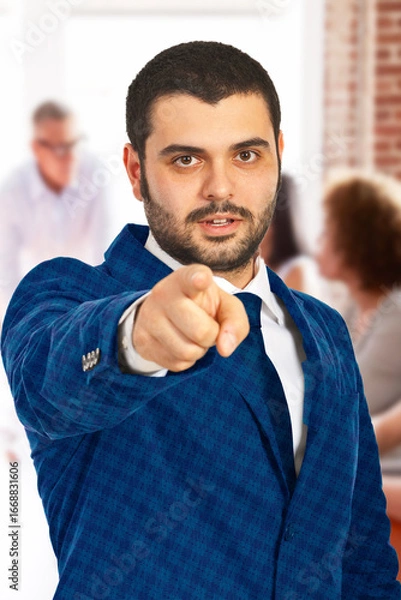 Obraz Confident young professional man in a blue suit pointing forward, focusing on business tasks in a corporate setting. Suitable for concepts like leadership, decision-making, and business communication.