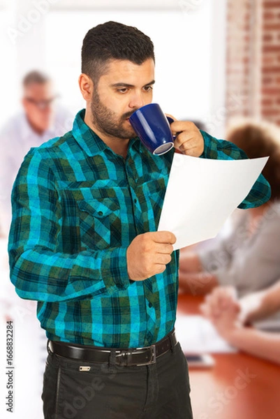 Obraz A man wearing a plaid shirt examines a document while enjoying a coffee in a workspace. Set in a modern, collaborative environment, it portrays focus, routine, and a professional atmosphere.