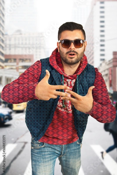 Fototapeta Young fashionable man stands on a city street, expressing cool and confident attitude. Shows modern urban style while conveying bold personality through striking hand gestures and stylish outfit.