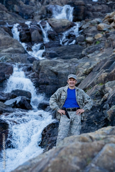 Fototapeta A contented Hunter in camouflage with a knife stands near a waterfall among stones