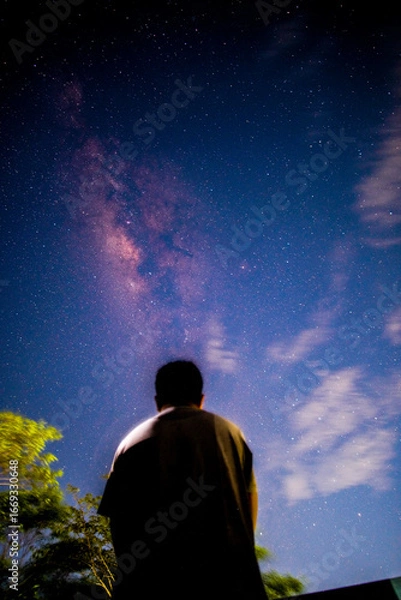 Fototapeta Back view of a person silhouetted against a beautiful night sky filled with stars and the vibrant Milky Way, evoking feelings of wonder and contemplation.