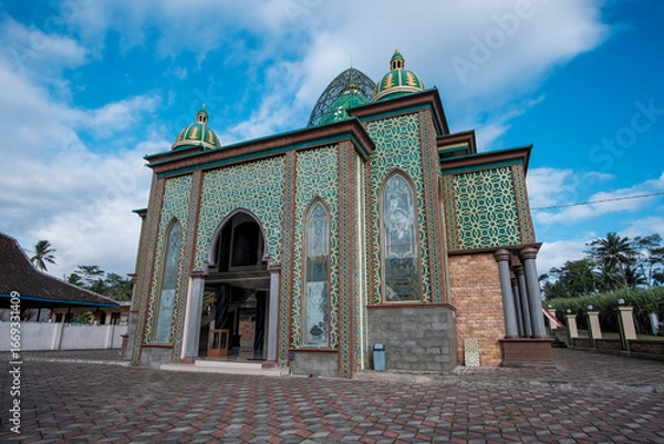 Fototapeta Beautiful Islamic mosque exterior featuring intricate gold geometric patterns on green walls, grand arched entrance, and multiple green domes under a blue sky.